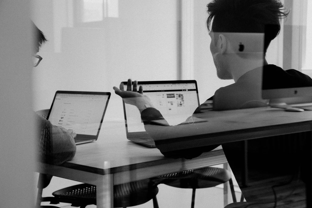 Black and white image of two people sat at their laptops. The photo appears to be taken through meeting room glass. The people are silhouetted and the right hand figure is gesturing at their laptop screen.